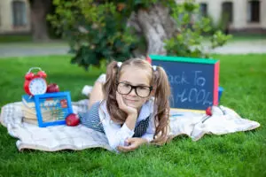 Cute smiling schoolgirl sitting on grass with lunch, books near the school. 