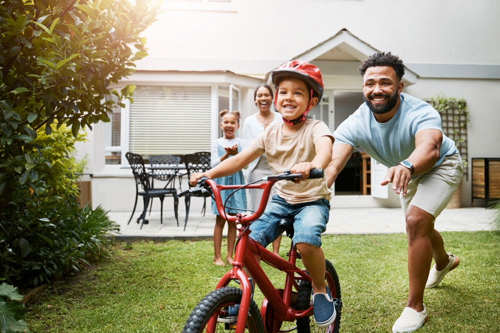 Learning, bicycle and proud dad teaching his young son to ride while wearing a helmet for safety in their family home garden. 